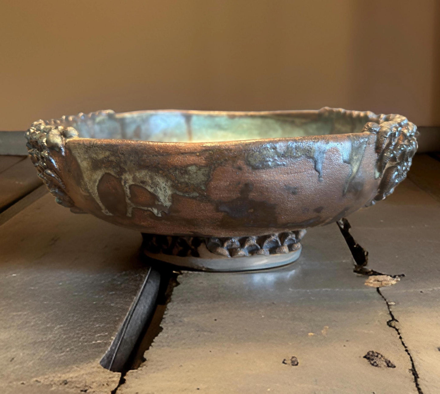 A ceramic vase for ikebana with a wide, shallow form and an ornate, textured rim. The  vase features a rustic finish with earthy tones of brown and green, highlighting its handmade quality. It rests on a circular base adorned with a beaded pattern.