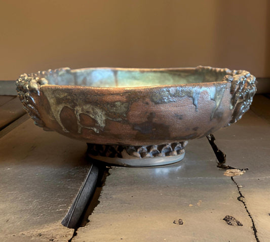 A ceramic vase for ikebana with a wide, shallow form and an ornate, textured rim. The  vase features a rustic finish with earthy tones of brown and green, highlighting its handmade quality. It rests on a circular base adorned with a beaded pattern.