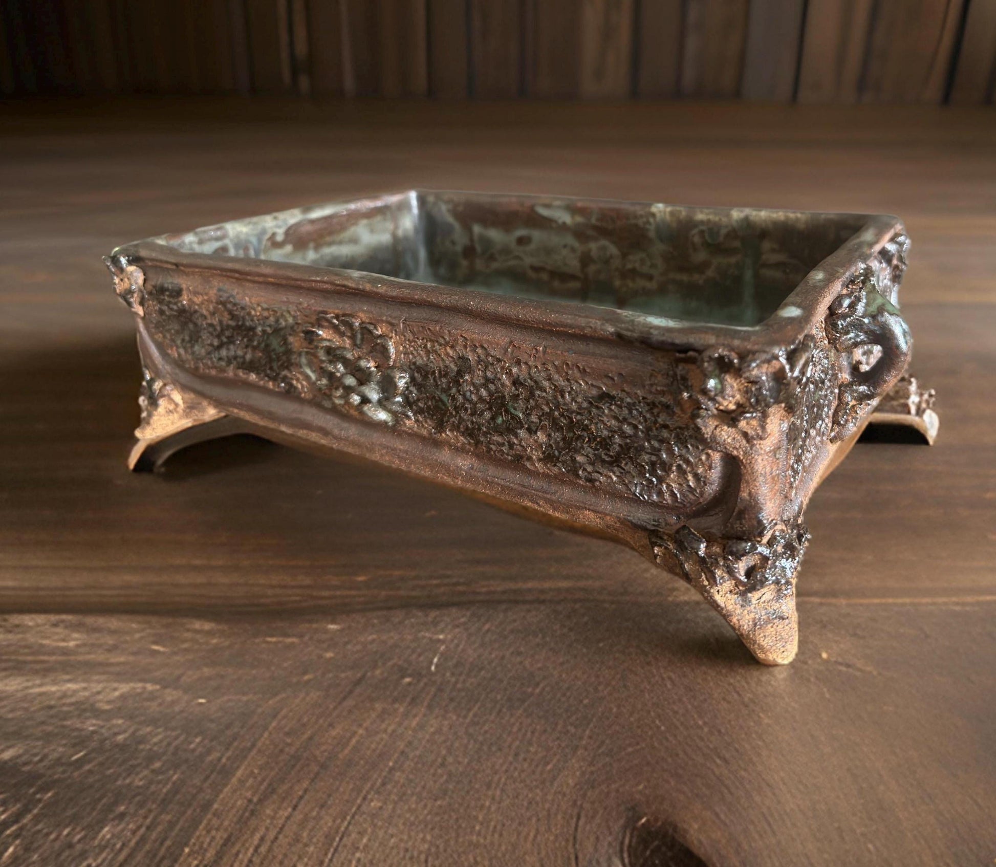A rectangular ceramic vase or container with noticeable texture and a metallic sheen, featuring supporting legs at each corner. The interior of the container is smooth, and overall it has an antique appearance, set against a wooden tabletop.