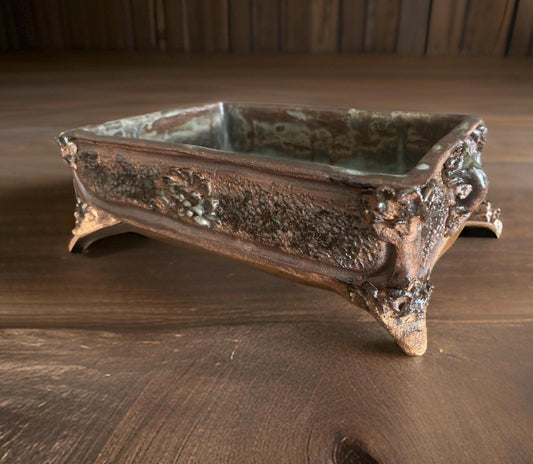 A rectangular ceramic vase or container with noticeable texture and a metallic sheen, featuring supporting legs at each corner. The interior of the container is smooth, and overall it has an antique appearance, set against a wooden tabletop.