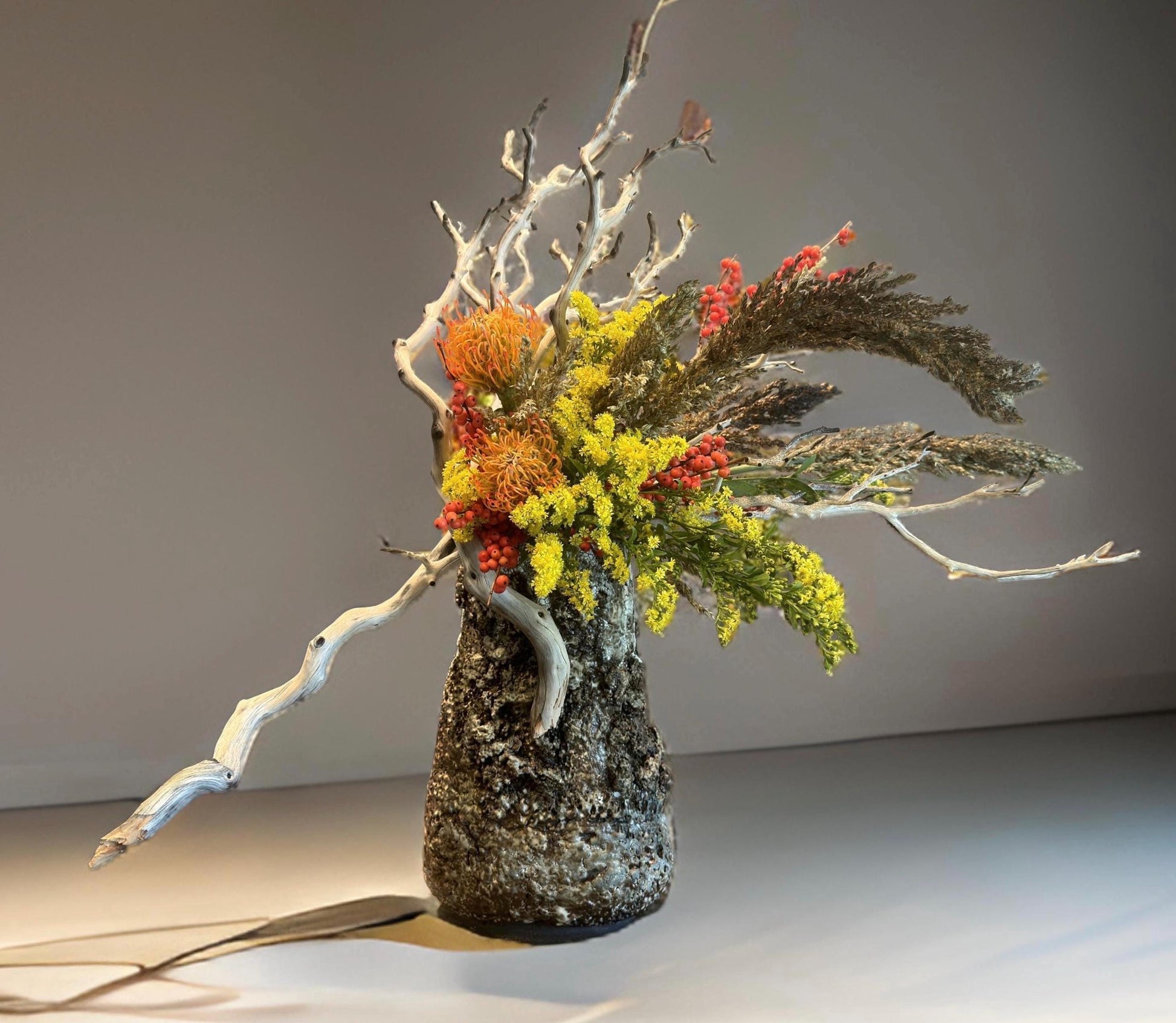 The red berries, and delicate grasses, showcased in a rustic, textured vase that resembles weathered stone. The vase has a unique, organic shape, tapering at the base, while its rough surface adds an earthy touch.