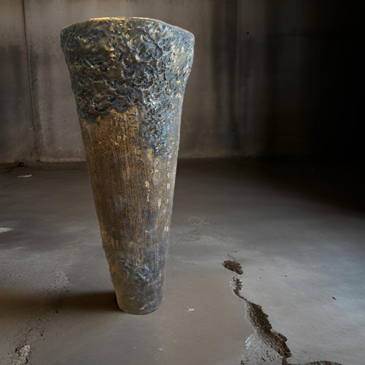 A tall, textured vase or sculpture made of dark, rough material with a gold speckled surface. It has a conical shape, wider at the top and narrowing towards the base, set against a minimalist gray concrete backdrop.