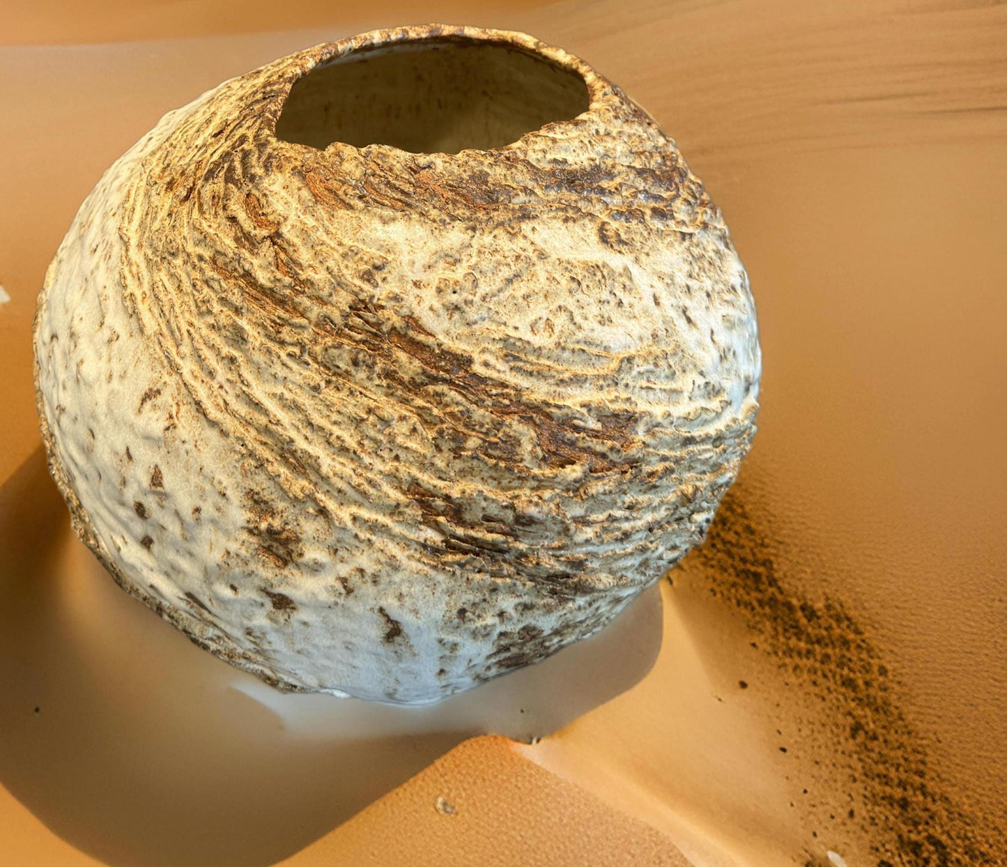 Hand made ceramic vase with earthy tones rests in a sandy desert landscape. The vase features an organic shape and an open top, making it a striking decorative piece. Its unique design captures attention and adds elegance to the natural surroundings.