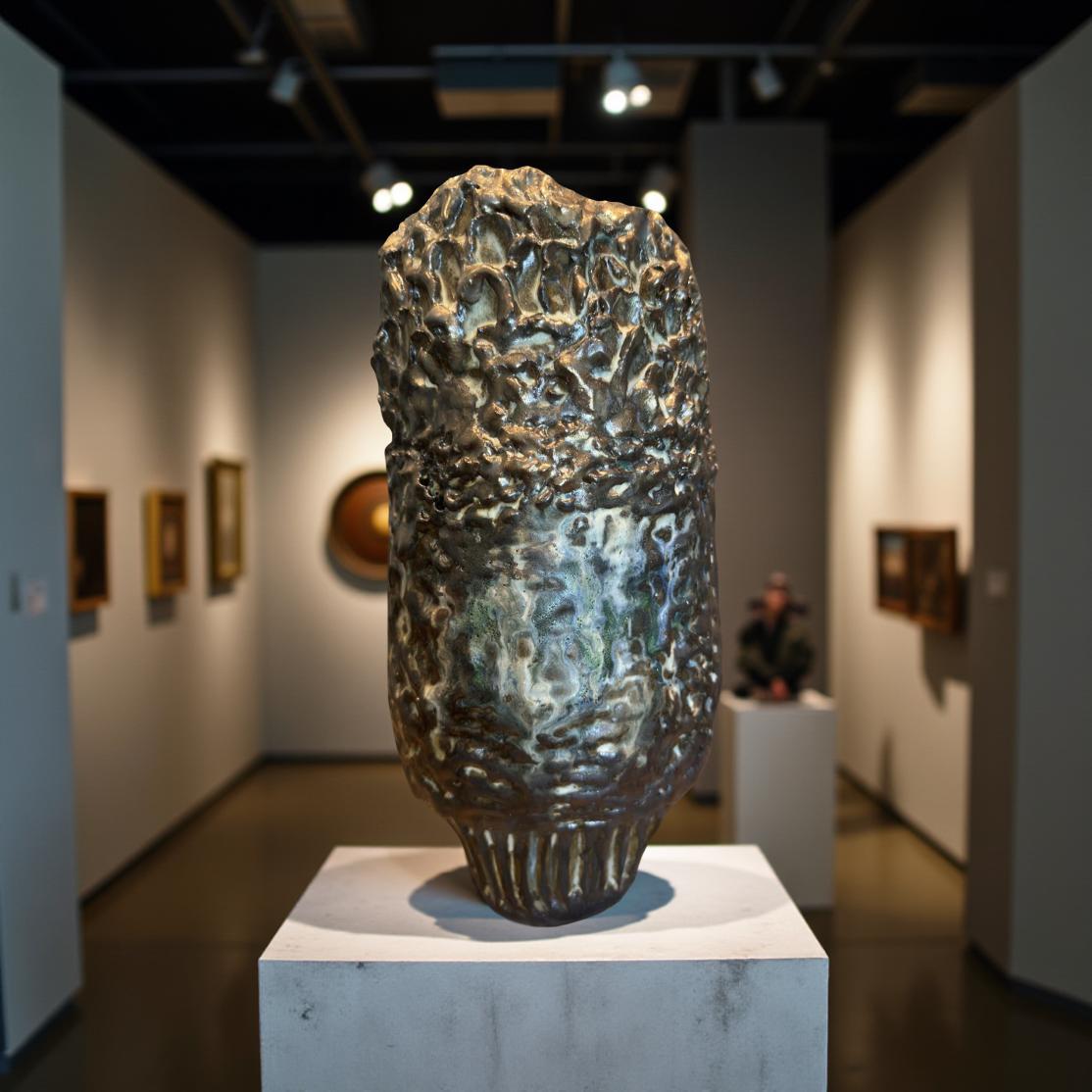 A dark gray and metallic ceramic vase with a textured, rock-like surface. The vase has an irregular, organic shape with a slightly bulbous body and a narrow base.  The surface is highly textured, with many small, raised bumps and crevices.  There are hints of green and off-white in the glaze, creating a mottled effect. The piece is a unique, artistic ceramic vessel.