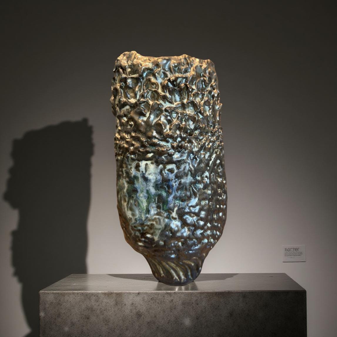 Dark metallic-green ceramic vase with a textured, rough surface. The vase has an irregular, organic shape, resembling a stylized flower or rock formation.  The glaze creates a three-dimensional effect, with peaks and valleys of color. The piece shows variations in shading and highlights, giving it depth and visual interest.  It is a unique, handcrafted ceramic art piece.