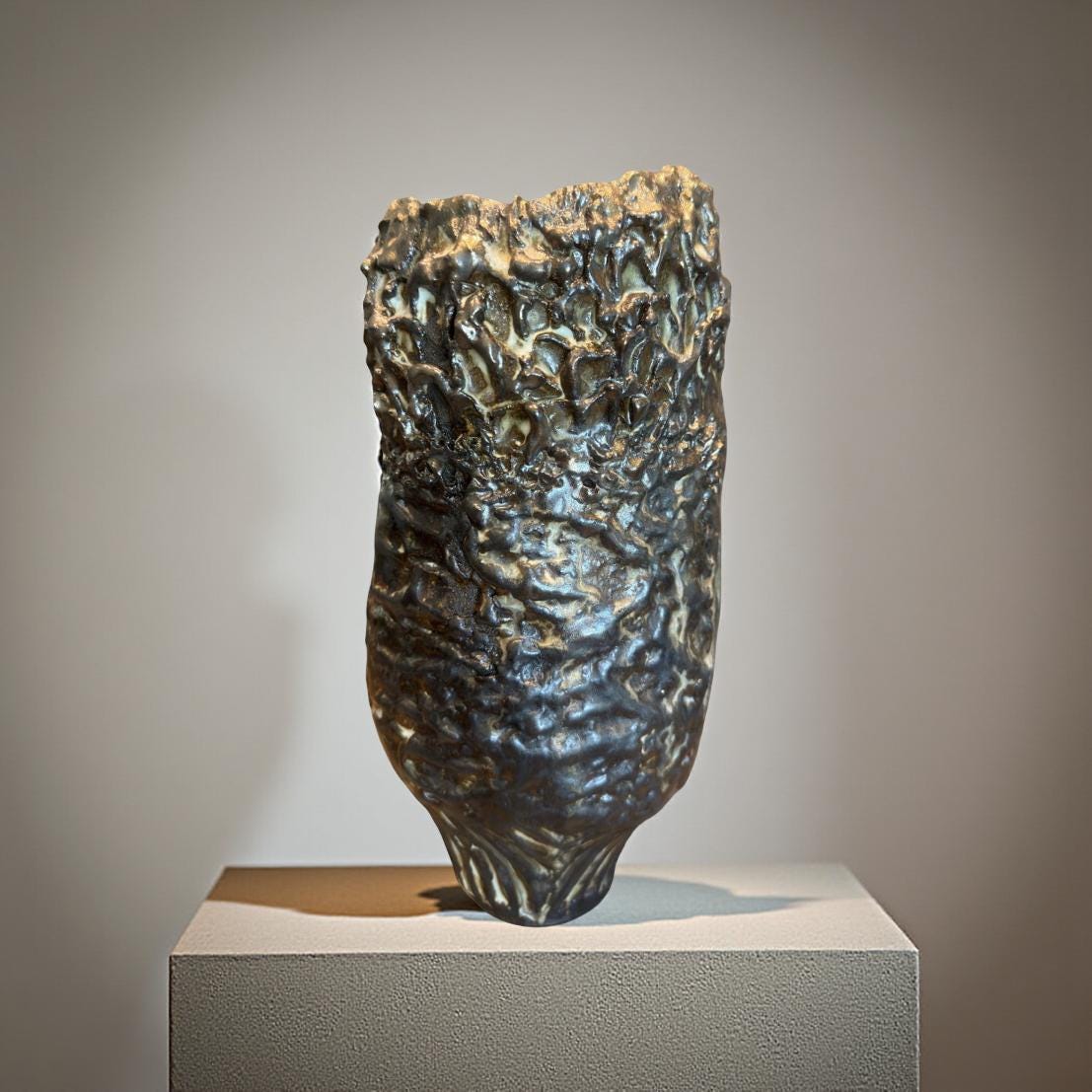 Close-up view of a dark gray ceramic vase with a textured, rough surface. The texture resembles a rocky or organic formation, with raised and uneven areas.  The vase has a slightly bulbous body that tapers towards a smaller base. The color is predominantly dark gray or black with hints of lighter gray or off-white in the crevices of the texture. The overall style suggests a rustic or artisanal aesthetic.