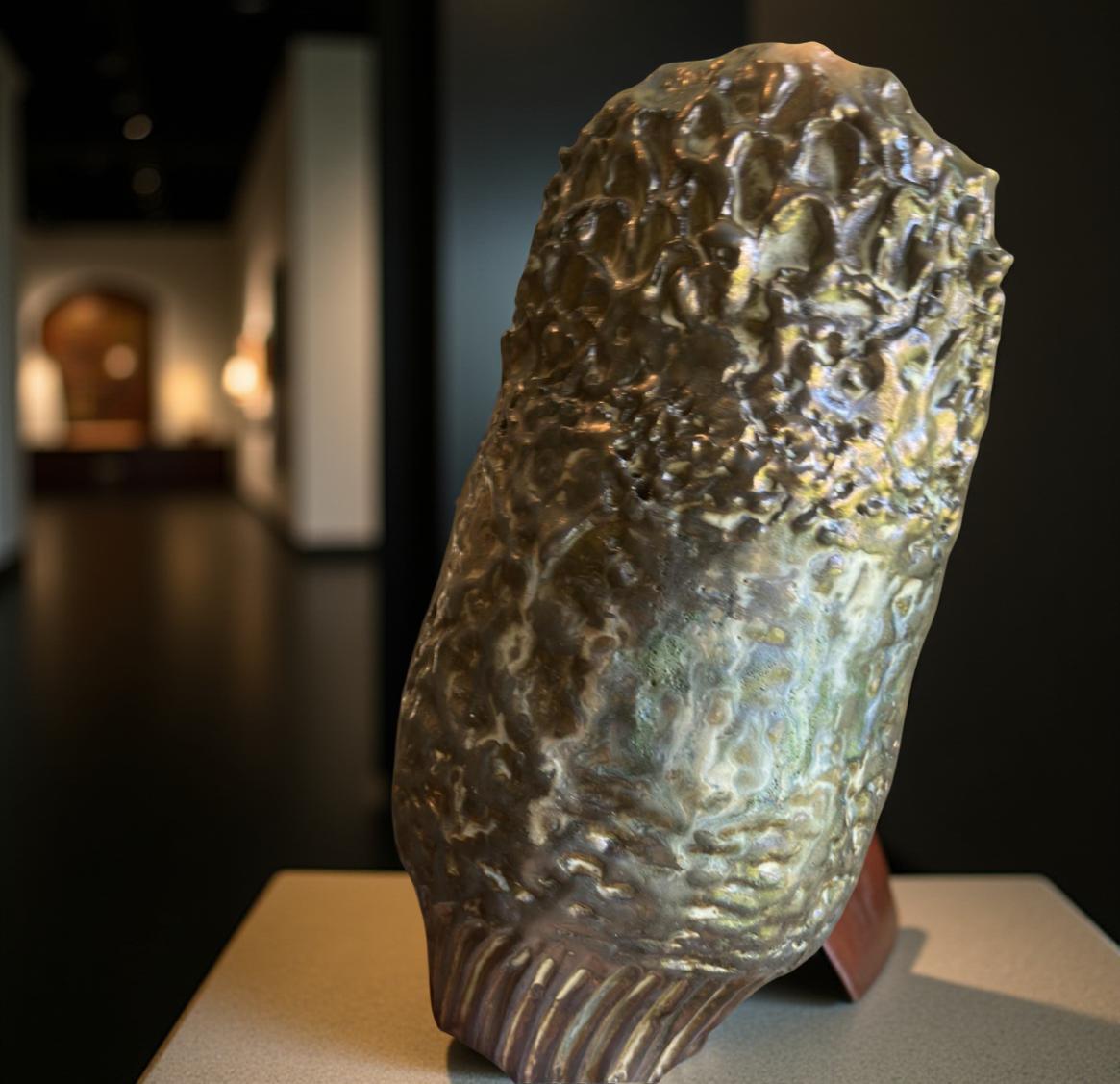 Close-up of a gold-toned ceramic sculpture resembling a stylized corn cob or pod. The surface is textured with numerous small, raised bumps, creating a rough, organic look. The color is a rich, metallic gold with subtle variations in tone and highlights. The base of the sculpture shows fine, vertical lines. The piece appears to be handcrafted and has a unique, artistic design.