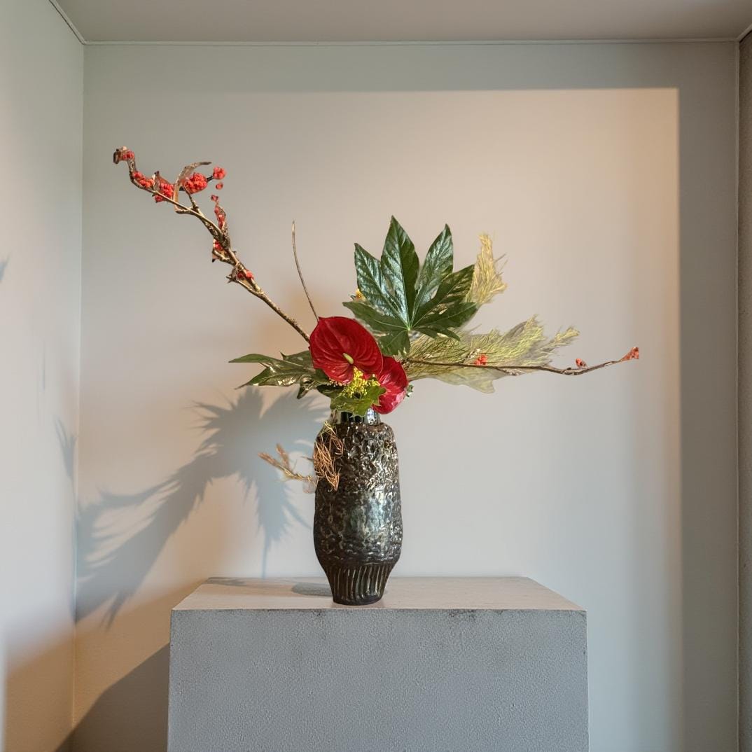 Ikebana flower arrangement in a dark gray textured ceramic vase.  Red Anthurium flowers, green leaves, tan branches, and dried beige grasses are artfully arranged. The vase is a dark mottled gray with a textured surface. The overall style is minimalist and modern.
