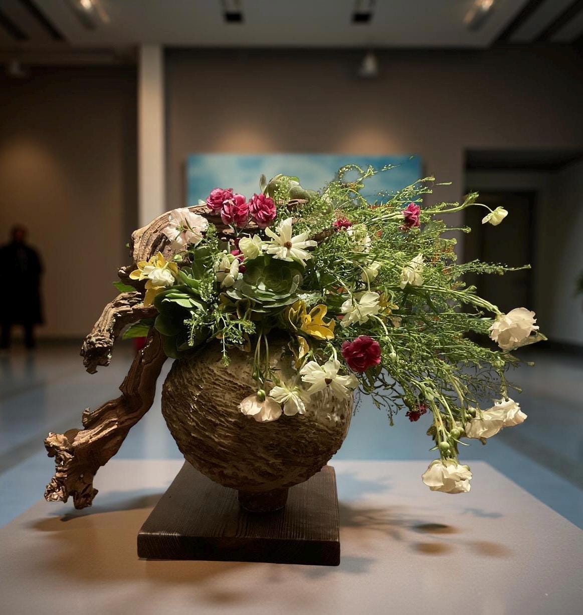 This floral sculpture was arranged in a ceramic vessel I hand-built and fired myself — rough, earthy, imperfect.
Driftwood curls from its side like a memory from the forest.
Every flower was placed by feel — not by rule.
White butterfly blooms, deep pink mini carnations, soft yellow alstroemeria, succulents, sweet peas…
All nestled in wild greenery